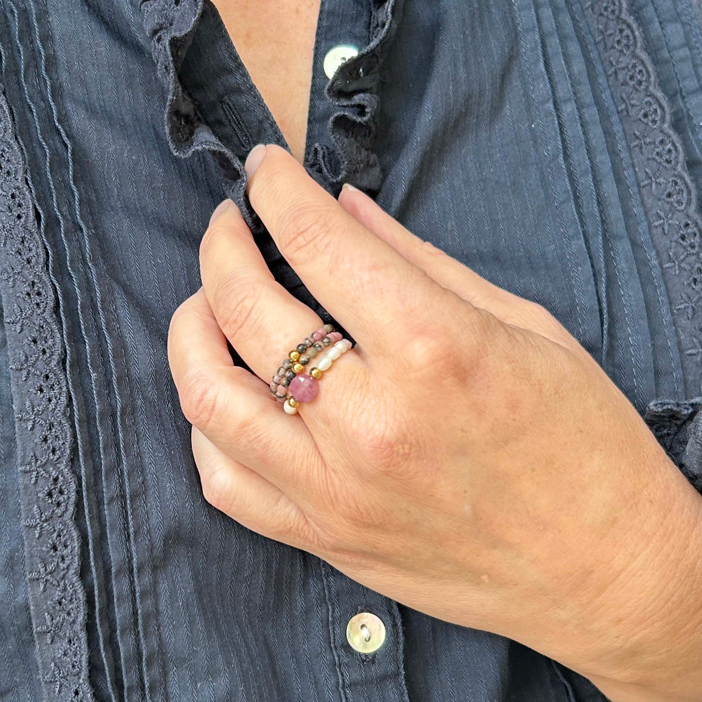 Close-up of a hand wearing a trio of delicate elastic rings with pink rhodonite gemstones and stainless steel gold beads. Feminine, affordable and stylish jewellery for everyday wear in the UK.
