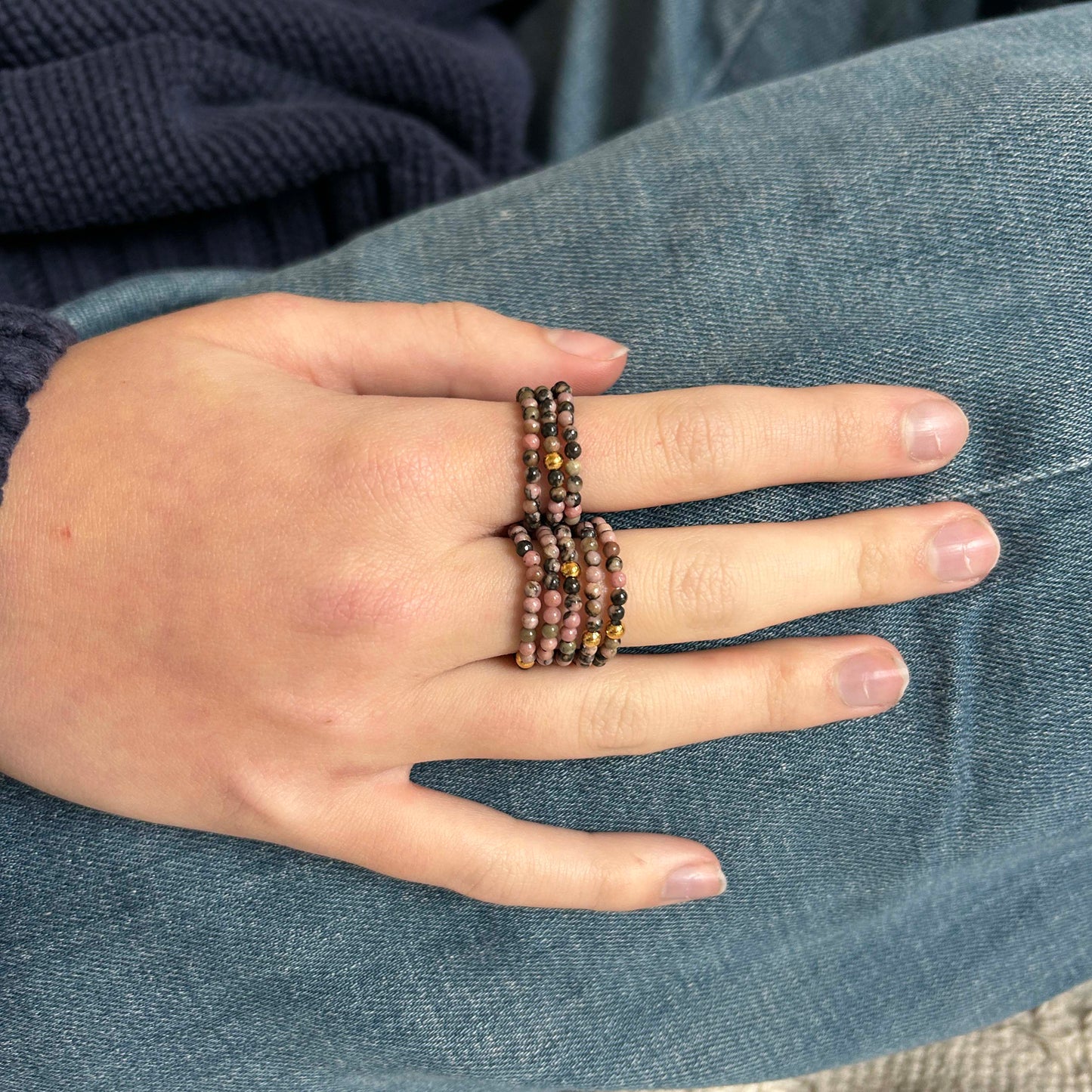 Stacked Leopoldine Aurora rings with rhodonite pink and gold beads, styled on hand over denim and navy knit.