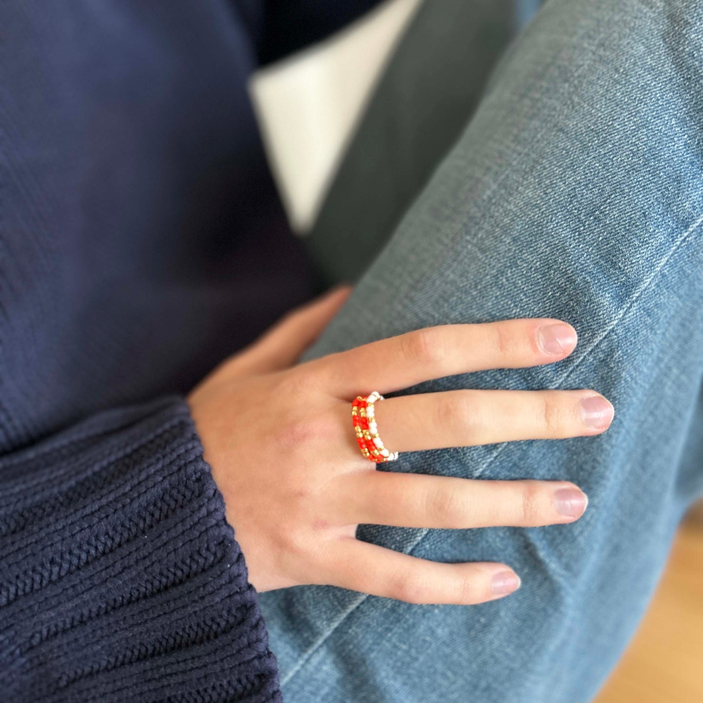 Affordable Leopoldine Confettis red beaded rings with gold details stacked on hand, styled with a cosy navy jumper and light denim jeans — vibrant and fun for everyday wear.