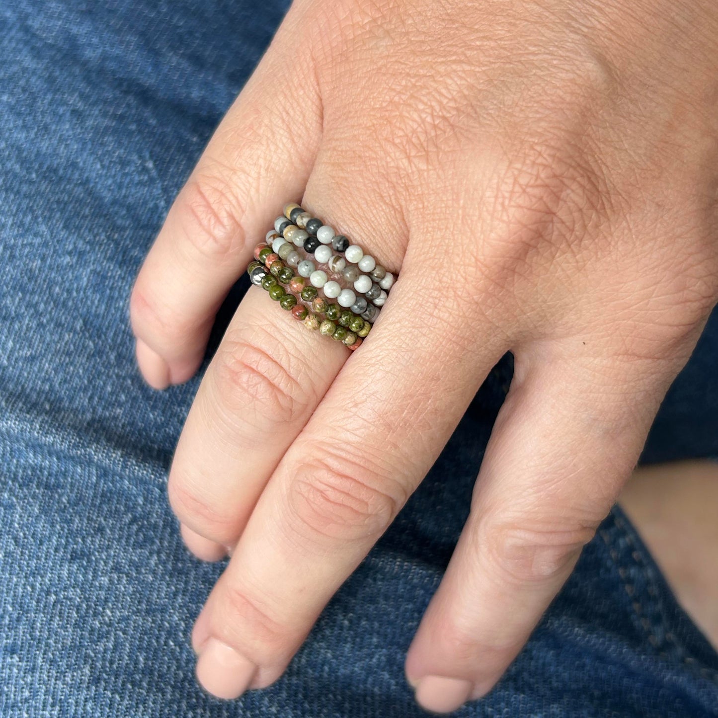 Stack of green and grey stone rings styled on hand with denim background, showcasing a moody natural palette.