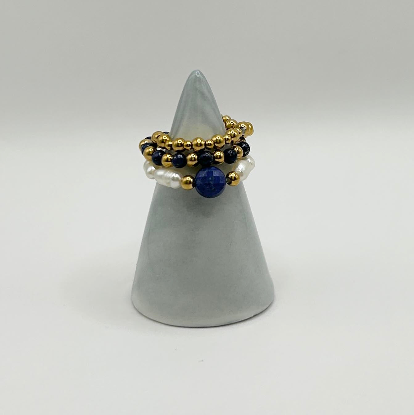 Leopoldine Amity ring trio with lapis lazuli, pearls and gold beads, styled on a sleek grey ring holder.