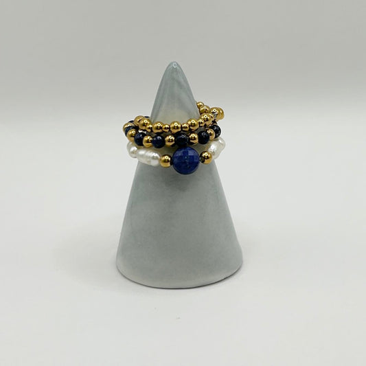 Leopoldine Amity ring trio with lapis lazuli, pearls and gold beads, styled on a sleek grey ring holder.