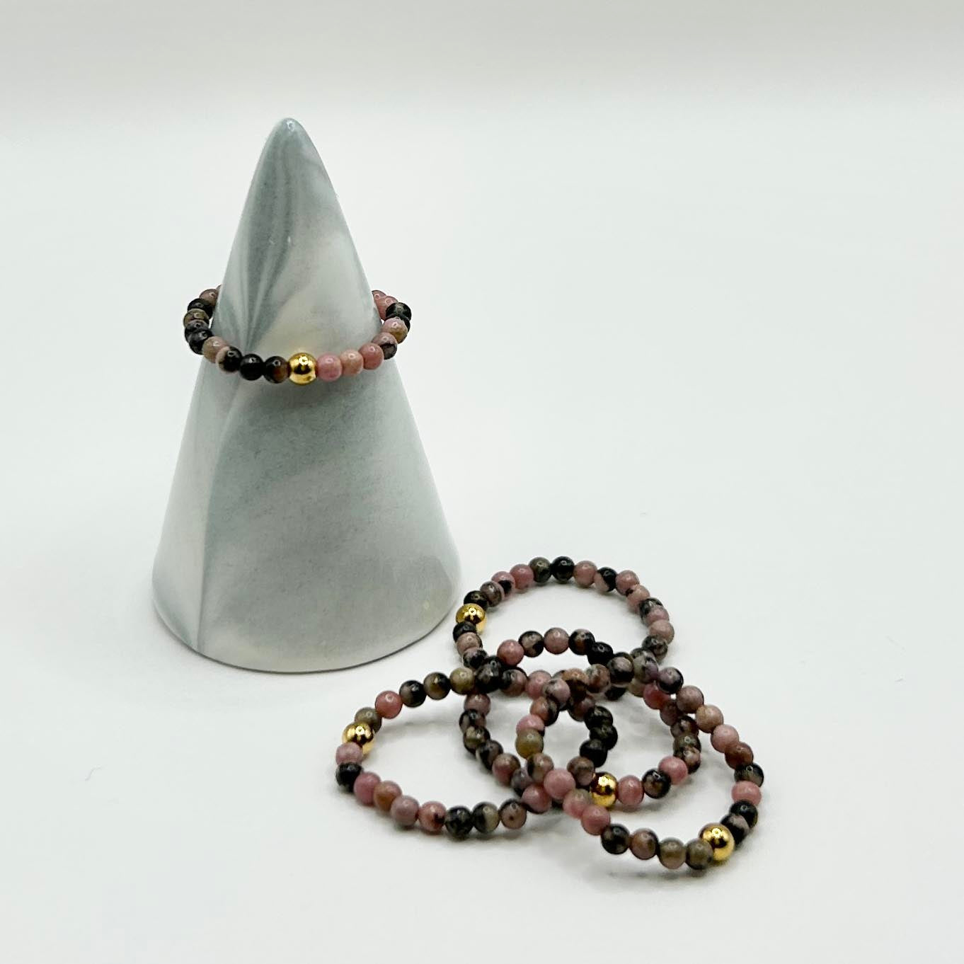 Set of Leopoldine stackable rings with pink rhodonite and gold beads, displayed on a cone stand with additional rings styled beside.