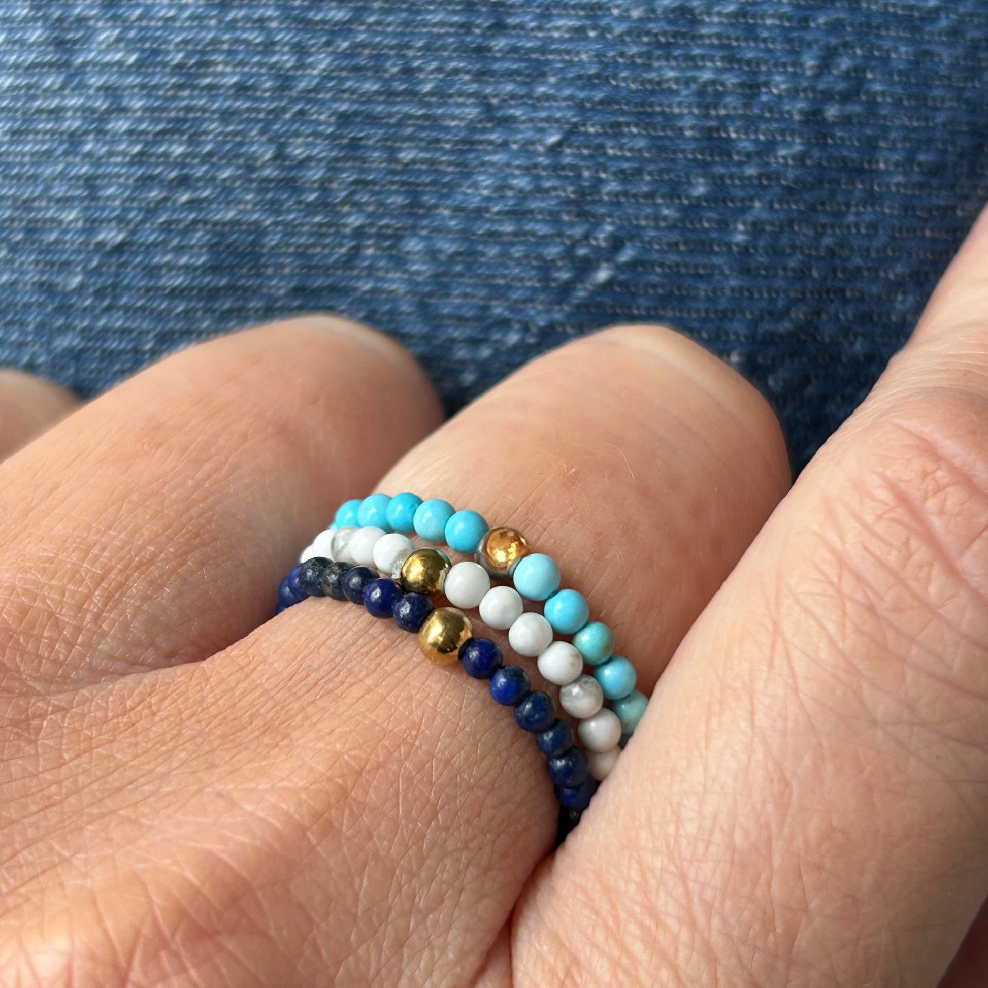 Close-up of a colourful ring trio in turquoise, white and navy, stacked on fingers with a denim background – stackable rings UK.